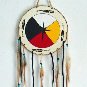 May include: A Native American dreamcatcher. The cream-coloured circular centre has a black, red, yellow, and white design. Black cords hang down, decorated with feathers and turquoise beads. The dreamcatcher is suspended by a tan cord.