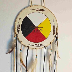 May include: A Native American medicine wheel dreamcatcher with a white, black, red, and yellow painted design. The dreamcatcher has a circle of brown leather with feathers hanging from it.