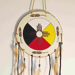 Könnte beinhalten: Ein Traumfänger aus weißem Leder mit einem bemalten indianischen Symbol in der Mitte. Das Symbol ist ein Kreis mit vier Farben: Schwarz, Rot, Gelb und Weiß. Der Traumfänger hat braune Lederbänder mit blauen Perlen und weißen Federn, die daran hängen.