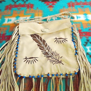 May include: A cream-coloured leather shoulder bag featuring a long braided strap and a flap closure with a single button. The front is decorated with a dark brown feather print and two sunburst motifs, accented by blue beads and long fringe.