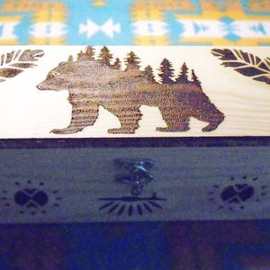 May include: A rectangular wooden box with a hinged lid. The lid features a laser-etched design of a bear with trees on its back, flanked by feather designs. The box has a metal clasp and decorative etchings.