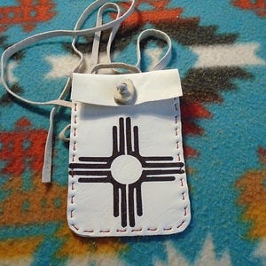 Peut inclure: Une pochette en cuir blanc avec des détails de couture rouge. La pochette a un rabat avec une fermeture à bouton et présente un symbole noir et blanc du symbole du soleil Zia.