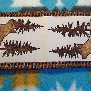 Puede incluir: Una cartera rectangular de cuero con un diseño temático de la naturaleza. La cartera presenta un panel central de color claro con un oso pardo y árboles grabados en la superficie. Los bordes son de color marrón oscuro con un borde cosido.