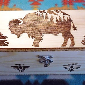 May include: A light-colored wooden box with a hinged lid and metal clasp. The lid features a detailed wood-burned design of a bison with mountains and trees, flanked by two feathers. The box has additional decorative elements.