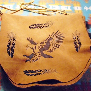 Könnte beinhalten: Eine hellbraune Ledertasche mit geflochtenem Riemen. Die Tasche hat ein schwarzes Adlerdesign mit ausgebreiteten Flügeln und vier schwarzen Federdesigns. Die Tasche ist mittelgroß und hat einen abgerundeten Boden.