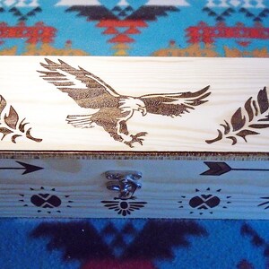 May include: A rectangular wooden box with a light finish, featuring etched designs. The lid displays an eagle and two feathers. The sides have arrows and sun symbols. A metal clasp secures the box.