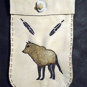 May include: Cream-colored leather pouch with a flap closure and a button. The pouch features a painted image of a brown animal and three black feathers. Red stitching outlines the edges.