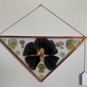 May include: A framed, diamond-shaped wall hanging with pressed flowers. The copper-colored frame holds a large, dark purple pansy, yellow and purple pansies, and small white flowers. A blank tag hangs from the bottom.
