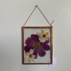 May include: A framed pressed flower art piece. The frame is a copper color, with a clear glass front. Inside are three pressed flowers: a large purple flower and two smaller white flowers. The art is hanging from a copper chain.