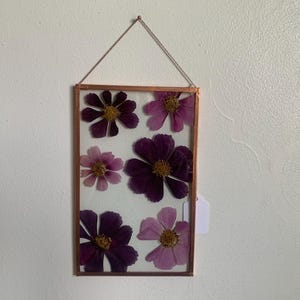 May include: A rectangular framed pressed flower art piece. The frame is copper-colored, and the flowers are various shades of purple and pink. The flowers are arranged on a white background and suspended by a chain.