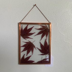 May include: A rectangular copper-framed glass hanging art piece. Inside, several deep red maple leaves are arranged against a clear background. The frame is suspended by a delicate chain, creating a simple yet elegant display.