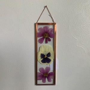 May include: A rectangular copper-framed pressed flower art piece. The artwork features three pressed flowers: two purple cosmos and a yellow and purple pansy. A delicate chain suspends the frame, showcasing the natural beauty of the botanical arrangement.