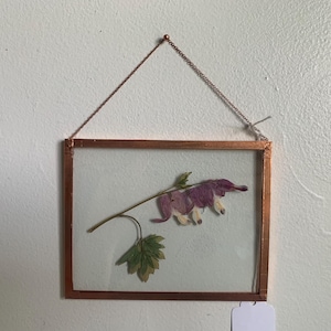 May include: A framed pressed flower art piece. The rectangular frame is copper-colored, with a delicate chain for hanging. Inside, a sprig of dried flowers and leaves in shades of purple, green, and white is displayed against a white background.