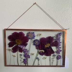 May include: A framed pressed flower art piece featuring deep purple cosmos flowers, lavender, and small white flowers. The flowers are arranged in a rectangular copper-colored frame, suspended by a delicate chain.
