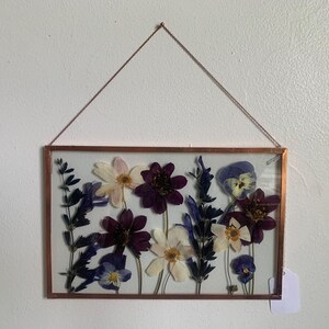 May include: A rectangular copper-framed pressed flower art piece. The artwork features an arrangement of dried flowers in shades of purple, white, and blue. The piece is suspended by a delicate copper chain, creating a unique wall hanging.