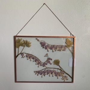 May include: A framed botanical art piece featuring pressed flowers. The copper-colored frame holds a light blue background with delicate purple and green floral arrangements. The artwork is suspended by a thin metal chain, creating a floating effect.