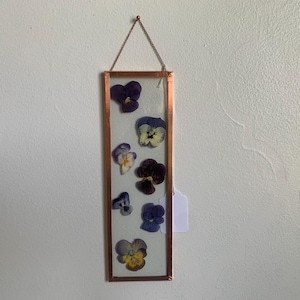 May include: A rectangular framed pressed flower art piece. The frame is copper-colored, and the flowers are various shades of purple, yellow, and white. The piece is suspended by a delicate chain.