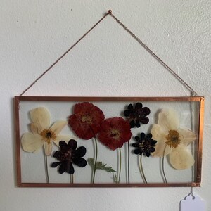 May include: A rectangular copper-framed wall hanging featuring pressed flowers. The arrangement includes red poppies, dark purple flowers, and cream-colored blooms. A delicate chain suspends the frame, showcasing a botanical theme.