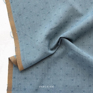 May include: Close-up of a blue fabric with a repeating cross pattern. The fabric has a textured appearance and a brown border. The word "FABLEISM" is printed in white at the bottom of the fabric.