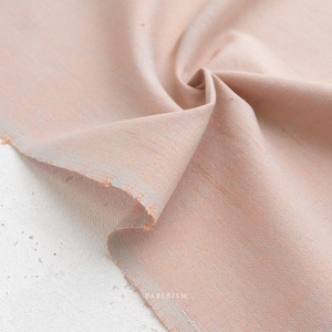 Pode incluir: Close-up de um tecido cor de pêssego com uma trama sutil e texturizada. O tecido está dobrado, revelando uma pitada de um tom azul claro ao longo das bordas. A borda bruta do tecido é visível, adicionando um toque de charme rústico.