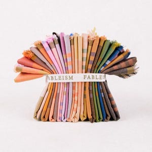 Puede incluir: Un conjunto de cuadrados de tela plegados de colores variados, abanicados y sujetos con una banda blanca. La tela presenta patrones de rayas. La banda dice "ABLEISM FABLE". Los bordes de la tela tienen un ribete de flecos.