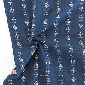 May include: Close-up of a navy blue fabric with a repeating pattern of white floral and orange cross designs. Vertical lines with small dashes separate the floral patterns. The word "FABLEISM" is printed in white.