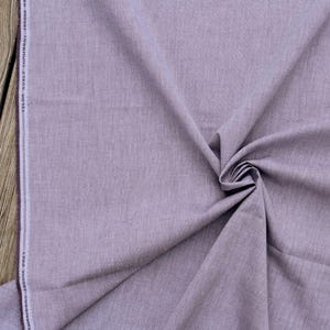May include: A close-up shot of a folded piece of fabric in a muted lavender color. The fabric has a textured appearance and is slightly creased, with a white stripe along one edge. The fabric is labeled "TAYLOR BUILT CHAMBRAY JEDDORE GREY."