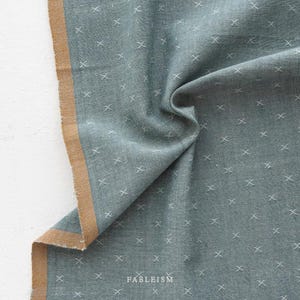 May include: A folded piece of teal fabric with a repeating white cross pattern. The edge of the fabric has a tan border. The word "FABLEISM" is printed in white at the bottom of the fabric. The fabric appears to be a linen or cotton blend.
