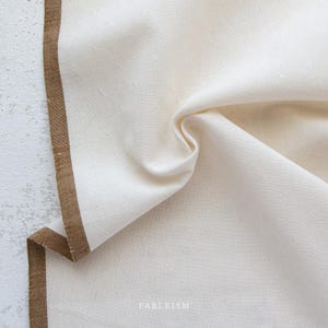 May include: A close-up of a white fabric with a brown border. The fabric is folded and has a textured surface. The text "FABLEISM" is printed on the fabric.