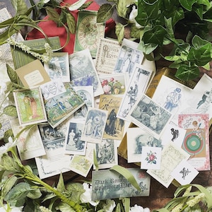 May include: A collection of vintage ephemera, including postcards and small cards, with illustrations of romantic scenes and figures. The items are arranged on a wooden surface and surrounded by white flowers and green foliage. The text "Dark Romance Ephemera Pack" is visible.