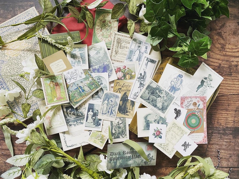 May include: A collection of vintage ephemera, including postcards and small cards, with illustrations of romantic scenes and figures. The items are arranged on a wooden surface and surrounded by white flowers and green foliage. The text "Dark Romance Ephemera Pack" is visible.