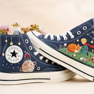 Può includere: Sneakers alte blu navy con ricami. Una scarpa presenta un pianeta con una rosa, l'altra una scena con una volpe, un ragazzo e stelle. Lacci e suole bianche. Il logo Converse è visibile.