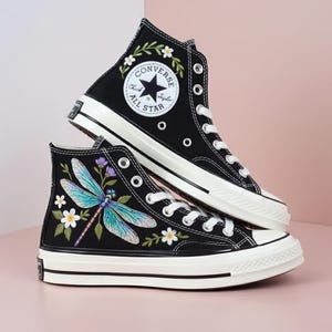 May include: Black high-top sneakers with white laces and soles. One shoe features a floral design around the Converse All Star logo, the other has a dragonfly and floral embroidery. The shoes have a classic, timeless design.