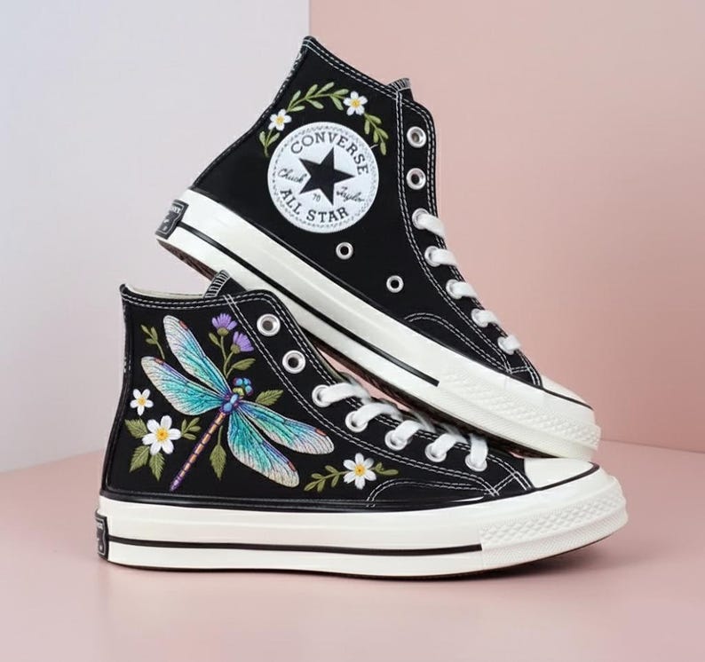 May include: Black high-top sneakers with white laces and soles. One shoe features a floral design around the Converse All Star logo, the other has a dragonfly and floral embroidery. The shoes have a classic, timeless design.