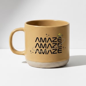 Könnte beinhalten: Eine beige Keramik-Tasse mit weißem Boden. Die Tasse zeigt das Wort "AMAZE" dreimal in Schwarz gestapelt, mit einer Planeten-Grafik und einem gelben geometrischen Design. Die Tasse hat einen gebogenen Henkel.