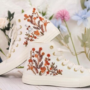 May include: Cream-colored high-top sneakers with intricate floral embroidery in shades of orange, brown, and silver. The sneakers feature gold eyelets and white laces. The design showcases a botanical theme.