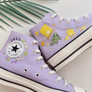Op de afbeelding: Een paar lavendelkleurige Converse All Star high-top sneakers. Eén schoen heeft een bloemenontwerp met de tekst "2025 Converse All Star". De andere heeft een geborduurd beeld van een gele lantaarn en een groene kameleon. Witte veters en zolen.