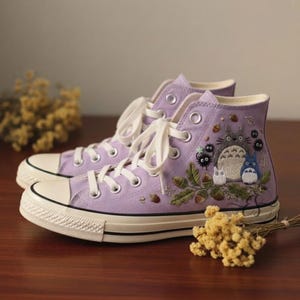 Op de afbeelding: Een paar hoge, lavendelkleurige canvas sneakers met witte veters en een witte rubberen zool. De sneakers hebben geborduurde afbeeldingen van stripfiguren, bladeren en eikels. De schoenen zijn lichtpaars.
