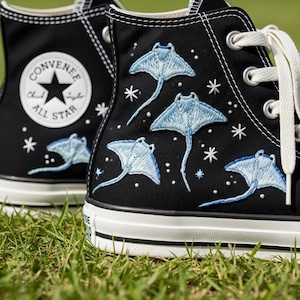 May include: Black high-top sneakers with embroidered light blue stingrays and white laces. The sneakers feature the Converse All Star logo on the side. The design includes white stitching and a white rubber sole.