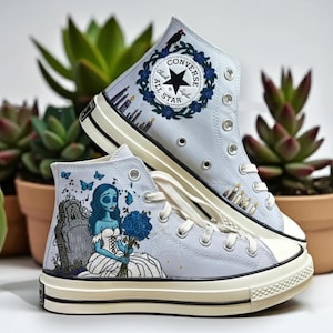 May include: Light blue high-top sneakers with custom artwork. One shoe features a woman holding blue flowers, a tombstone, and butterflies. The other shoe has a floral wreath with a black bird and the Converse logo. The sneakers have white laces and soles.