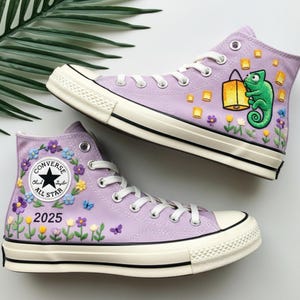 Op de afbeelding: Een paar lavendelkleurige high-top sneakers met witte veters en zolen. De sneakers zijn geborduurd met bloemmotieven, vlinders en een cartoonkameleon met een lantaarn. De tekst "2025" is ook op één schoen geborduurd.