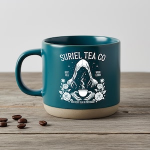 Op de afbeelding: Teal keramische mok met de tekst "Suriel Tea Co" en een witte afbeelding van een figuur met capuchon die een theekopje vasthoudt, omringd door rozen. De mok heeft een beige basis en een donker teal handvat.