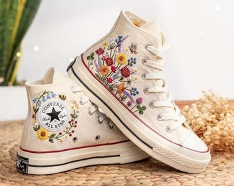 May include: Cream-colored high-top sneakers with floral embroidery. The sneakers feature a black star logo and a red stripe along the sole. The embroidered flowers include red, yellow, blue, and purple.