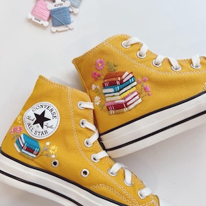 Può includere: Sneakers alte giallo senape con lacci e suole bianche. Le sneakers presentano ricami di pile di libri e motivi floreali. Il logo Converse è visibile sul lato. Un approccio unico e artistico alle calzature classiche.
