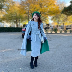 May include: A light gray dress with a matching cape and a green lining. The outfit includes a green beret, black gloves, and black boots. A small silver clutch is held in the hand. The background features trees with yellow leaves.