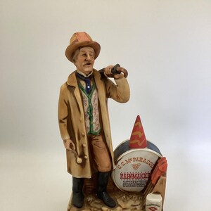 May include: A porcelain figurine of a man in a brown coat and hat holding an umbrella, standing next to a drum with the text "C.C. Mc. Parasol Rainmaker Satisfaction Guaranteed".