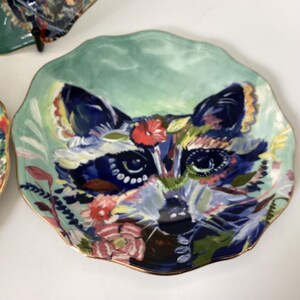May include: A decorative ceramic plate with a raccoon design. The plate features a light green background and a detailed raccoon face with floral accents in shades of blue, red, and yellow. The rim is trimmed with gold.