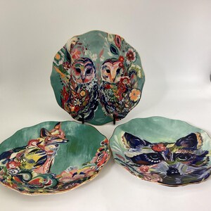 May include: Three decorative ceramic plates with a teal background and gold trim. The plates feature colorful illustrations of an owl pair, a fox, and a raccoon. The owl plate is the largest, with the fox and raccoon plates being smaller.