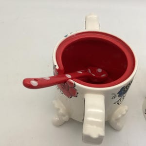 May include: A white ceramic sugar bowl with a red interior and a matching spoon. The bowl features floral designs in pink and blue, with a red rim and a red spoon with white polka dots. The bowl has a handle and rests on three small feet.