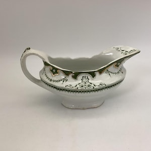 May include: White ceramic gravy boat with a scalloped rim and a handle. The gravy boat is decorated with green and gold floral patterns. The design is repeated around the base and rim of the boat. The gravy boat is a classic piece of tableware.